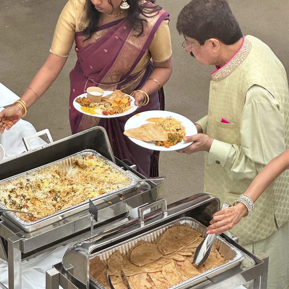 Two guests in traditional Indian attire serve plates of biryani, vegetables, curry, and chapati from a buffet.