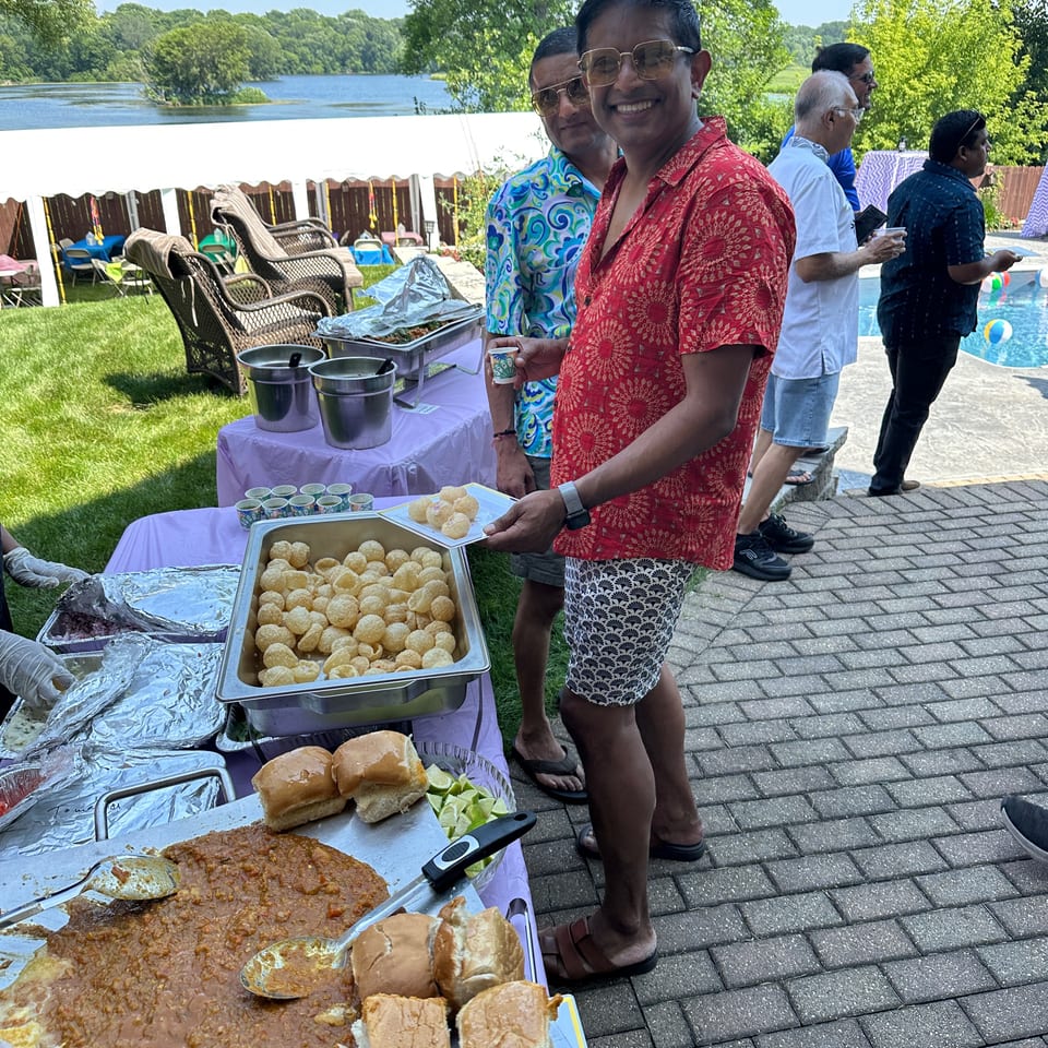 Guests gather around an outdoor Indian food buffet beside a pool and lake, with pani puri, curry, bread, and serving trays.