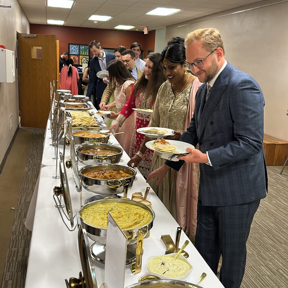 Guests in formal and traditional attire serve themselves from a buffet of Indian dishes in a banquet room.