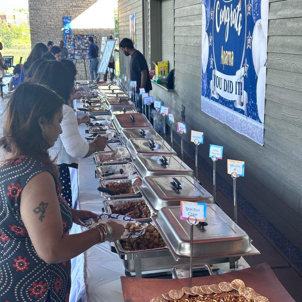 Guests serve themselves Indian dishes from chafing trays at an outdoor buffet beneath a “You Did It!” celebration banner.