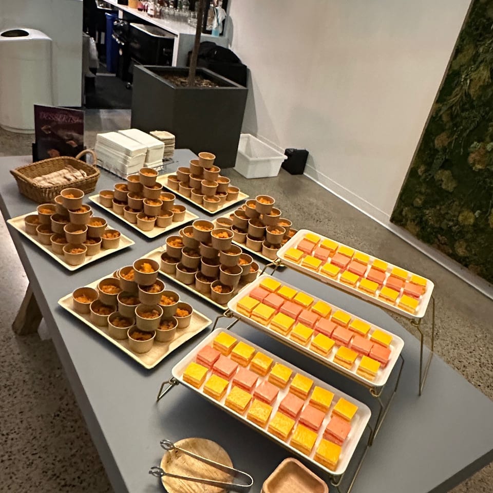 Buffet table displaying individual savory bites and colorful layered desserts in a modern restaurant beside a bar and indoor tree.