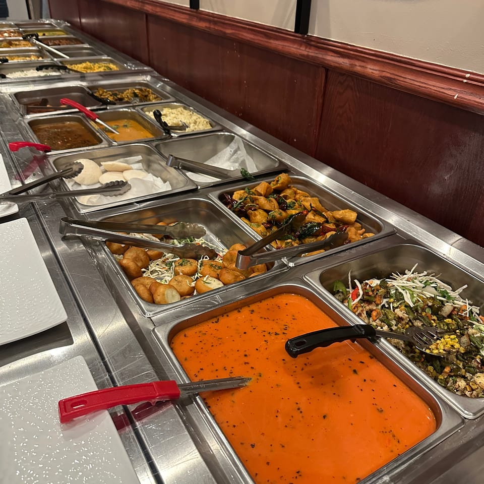 Buffet station displaying assorted curries, fried appetizers, vegetables, rice, sauces, and serving tongs in stainless steel trays.