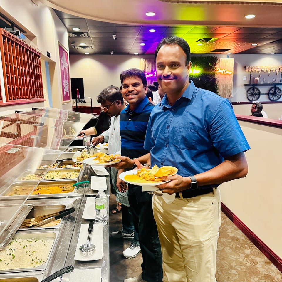 Smiling diners hold plates beside a buffet of Indian dishes in a warmly lit restaurant.
