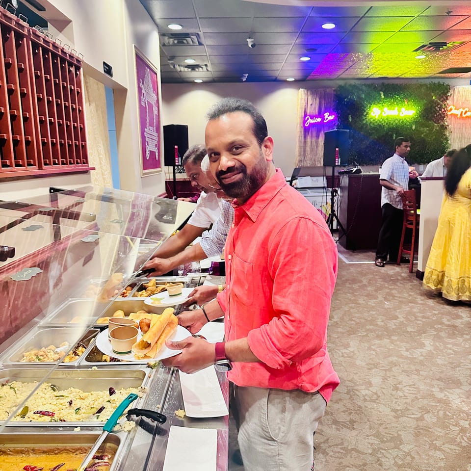 Man in a coral shirt serves himself food from a buffet counter inside a colorful restaurant.