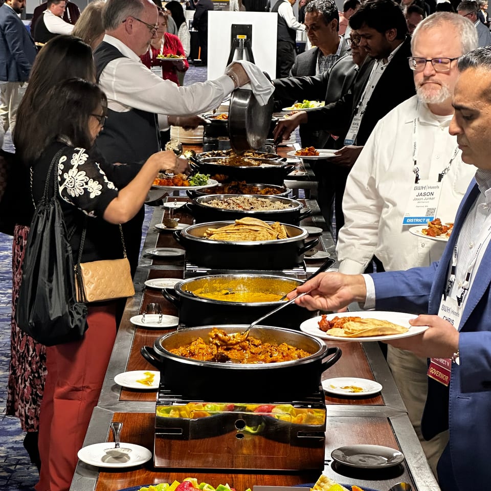 Crowded buffet featuring Indian dishes, rice, curry, naan, and colorful snacks as guests serve themselves at an event.