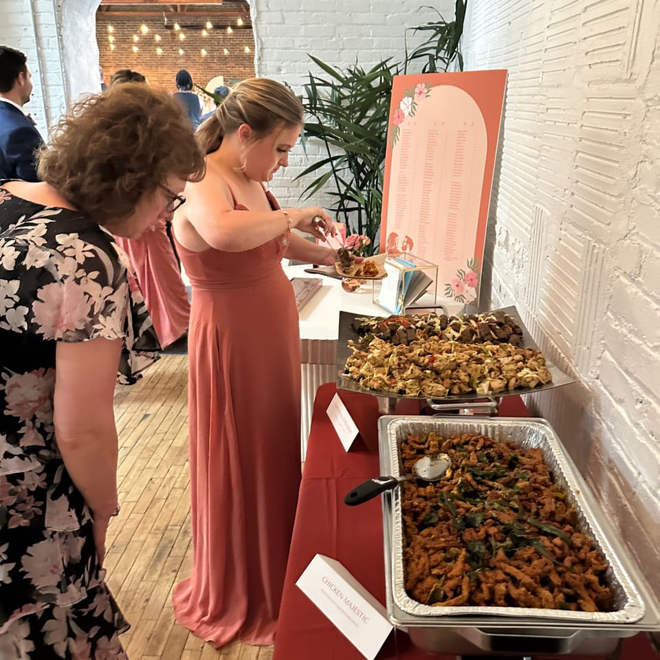 Guests browse a buffet featuring trays of chicken dishes beside a floral menu sign in a warmly lit restaurant.
