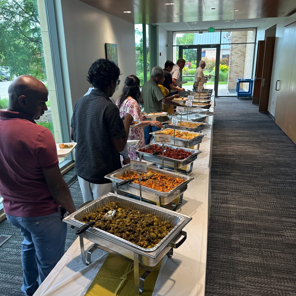 People serve themselves Indian dishes from a buffet line in a bright restaurant hallway.