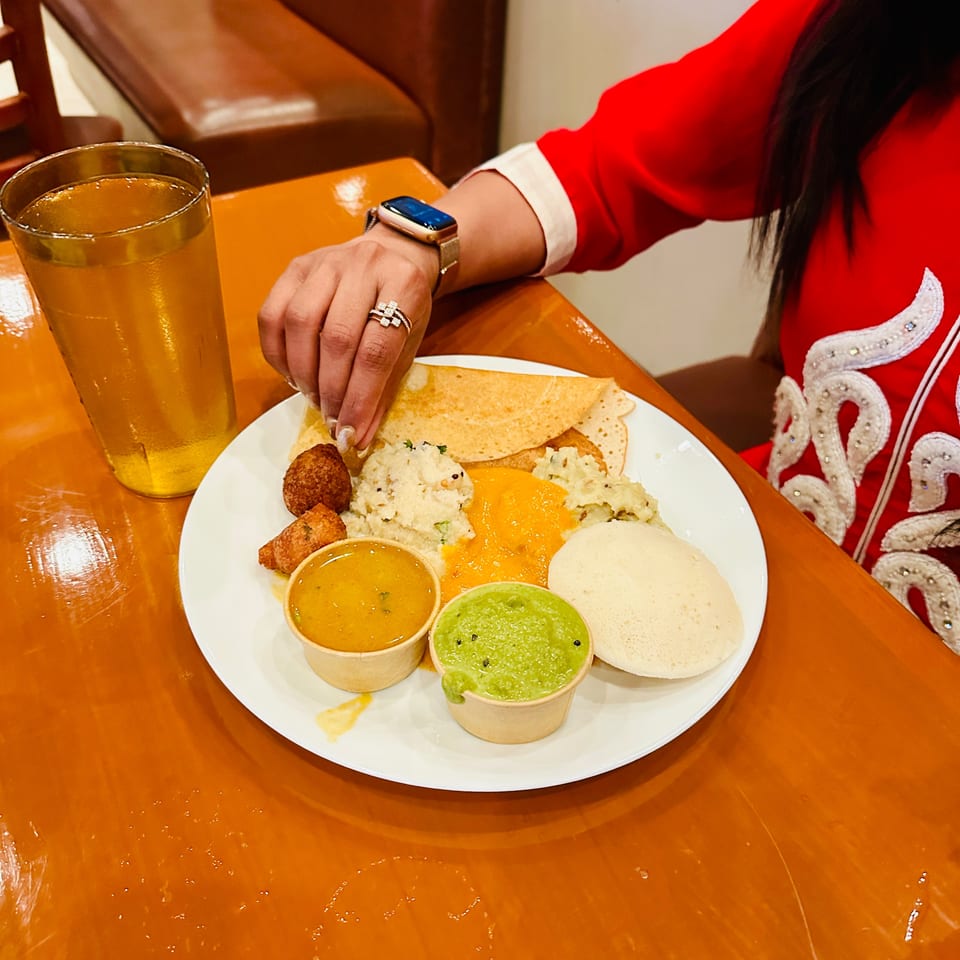 Diners enjoy a plate of flatbreads, rice, fritters, sauces, and creamy sides beside a glass of orange drink.