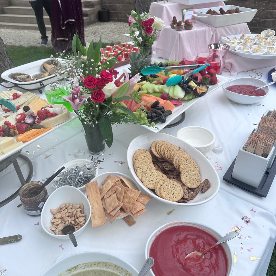 Outdoor buffet table displaying fruit, cheese, crackers, dips, pastries, fried appetizers, and floral arrangements.