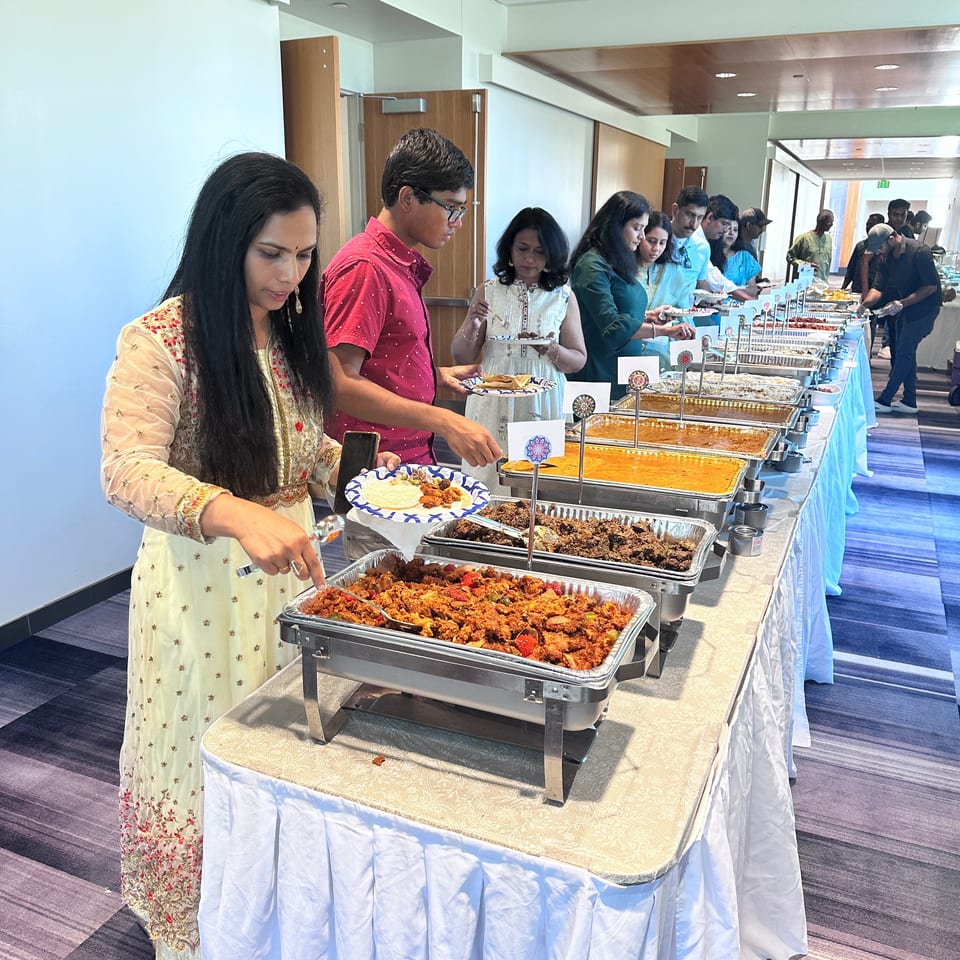 Guests serve themselves Indian dishes from a long buffet table in a brightly lit banquet room.