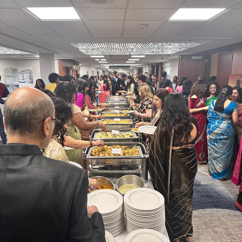 Crowded indoor Indian buffet with guests serving food from chafing dishes, surrounded by stacks of plates.