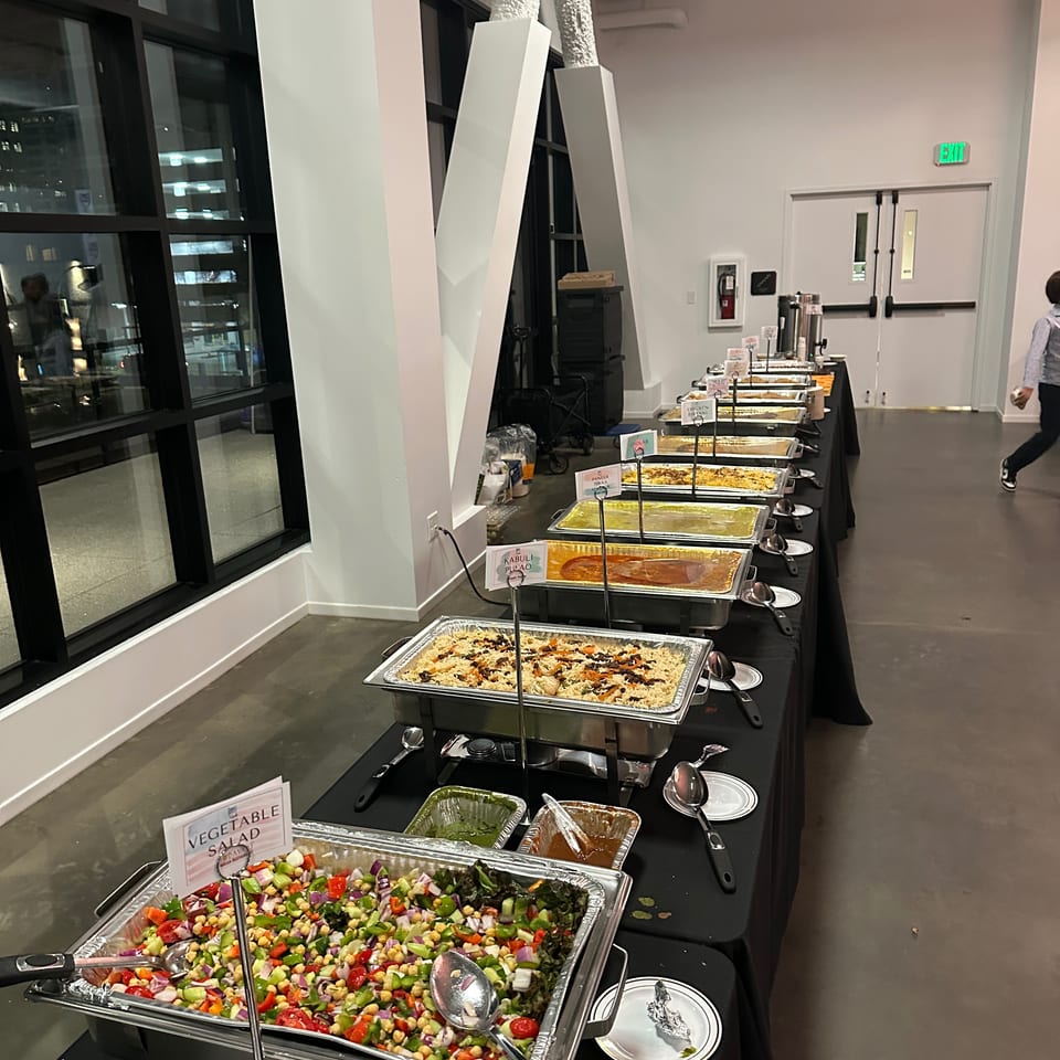 Buffet table displays Indian and Middle Eastern dishes, including biryani, paneer tikka, kabuli pulao, sauces, and vegetable salad.