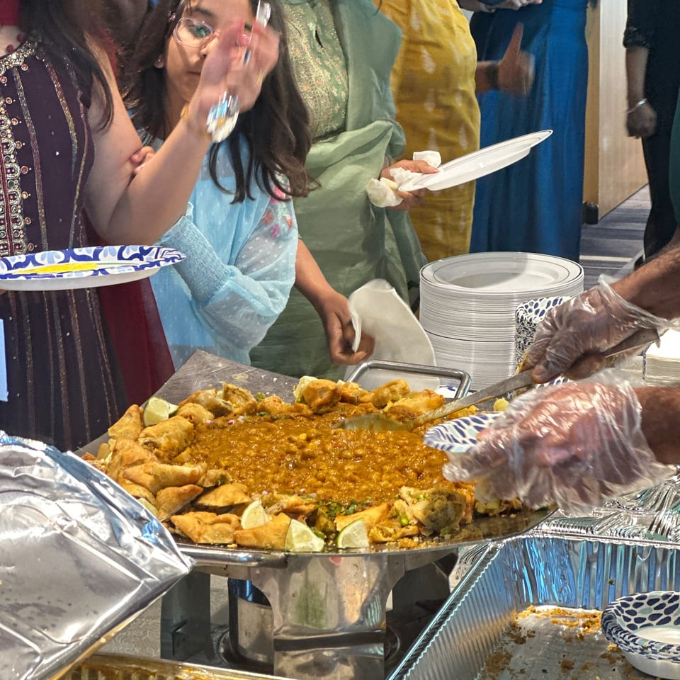 Guests serve themselves from a buffet featuring Indian dishes, fried appetizers, lime wedges, and stacks of plates.