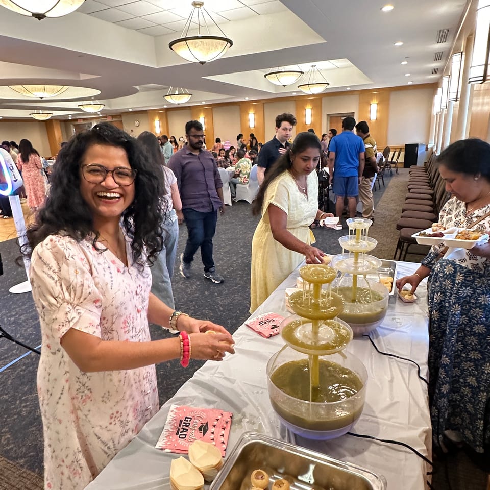Busy restaurant gathering with guests serving Indian snacks and sauces from a tiered fountain buffet beneath decorative chandeliers.