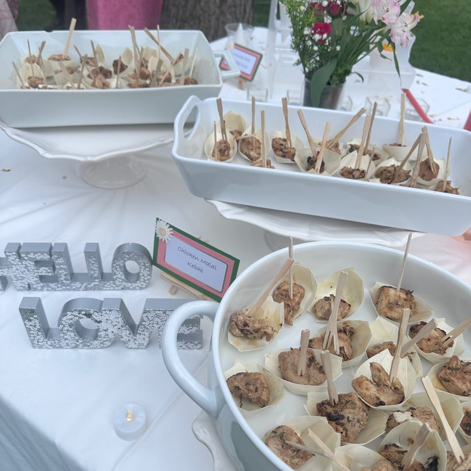 Outdoor buffet table displaying skewered chicken malai kebabs, small cups of snacks, and a flower arrangement.